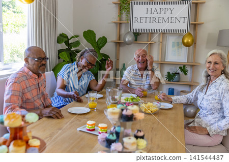 Diverse senior friends celebrating retirement in dining room at table with banner, balloons Diverse senior friends celebrating retirement in dining room at table with banner, balloons 141544487