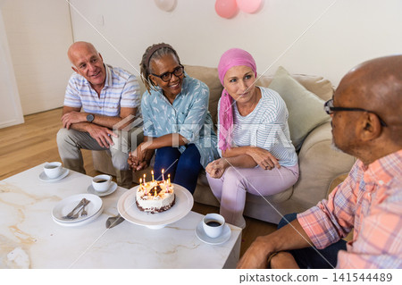 Diverse senior friends celebrating birthday at home around coffee table with cake, balloons Diverse senior friends celebrating birthday at home around coffee table with cake, balloons 141544489