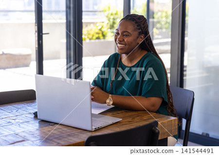 African american woman working at table in office using laptop, reviewing paper, copy space 141544491