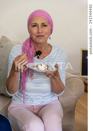 Senior female wearing headscarf sitting on sofa at home holding fork over cake on plate Senior female wearing headscarf sitting on sofa at home holding fork over cake on plate 141544492