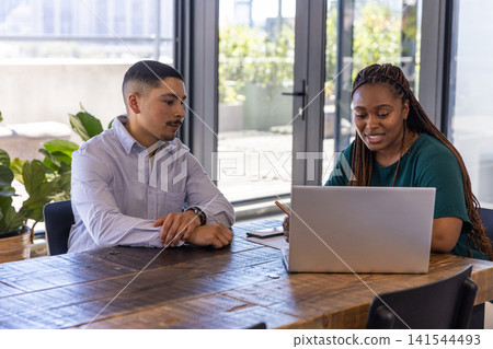 Diverse coworkers sitting at table in office reviewing notebook with pencil next to laptop 141544493