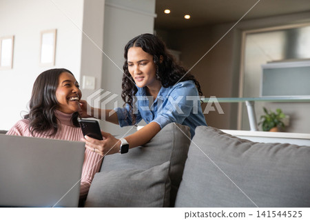 Diverse female friends sitting on gray couch sharing laptop and smartphone in living room Diverse female friends sitting on gray couch sharing laptop and smartphone in living room 141544525
