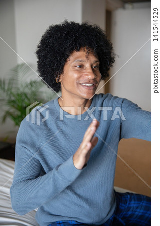 African american man gesturing, smiling while sitting on bed in bedroom with potted plant African american man gesturing, smiling while sitting on bed in bedroom with potted plant 141544529