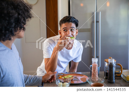 Diverse male friends eating avocado toast and talking at home kitchen island with French press Diverse male friends eating avocado toast and talking at home kitchen island with French press 141544535