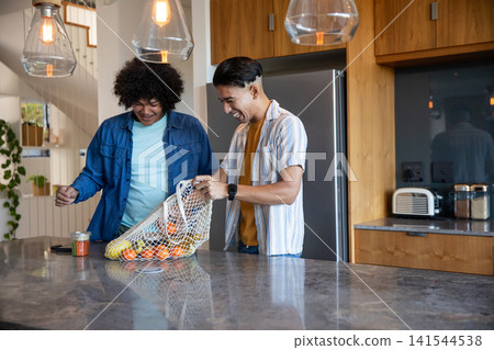 Diverse male friends unpacking mesh bag of oranges and apples on marble island in modern kitchen Diverse male friends unpacking mesh bag of oranges and apples on marble island in modern kitchen 141544538