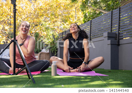 Diverse female friends practicing yoga in sportswear on yoga mats in backyard recording Diverse female friends practicing yoga in sportswear on yoga mats in backyard recording 141544540