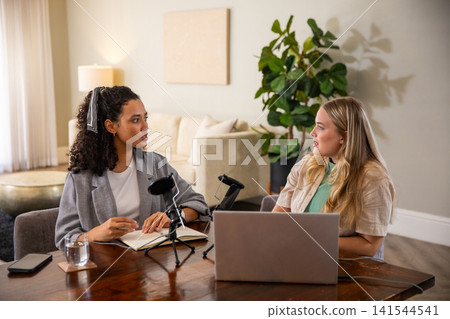 Female coworkers recording discussion using microphones and typing on laptop in living room Female coworkers recording discussion using microphones and typing on laptop in living room 141544541