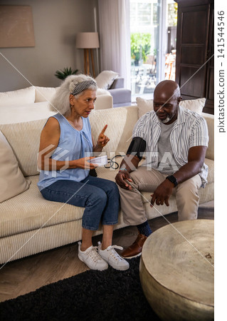 Diverse senior couple checking blood pressure with digital cuff monitor on sofa in living room Diverse senior couple checking blood pressure with digital cuff monitor on sofa in living room 141544546