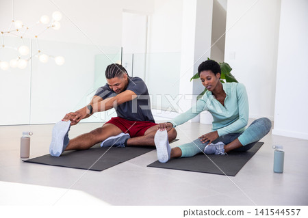 Diverse workout partners stretching hamstrings on black mats in living room with water bottles Diverse workout partners stretching hamstrings on black mats in living room with water bottles 141544557