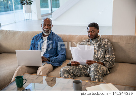 African american father and son reviewing papers on couch in living room with laptop, notebook African american father and son reviewing papers on couch in living room with laptop, notebook 141544559