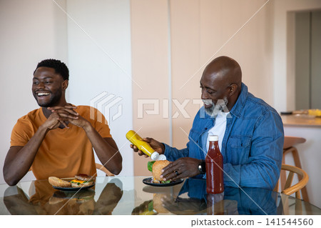 African american father and son at glass table at home eating burgers with mustard, ketchup 141544560
