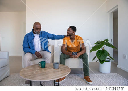 African american father and son on chairs in living room sharing smartphone by coffee table African american father and son on chairs in living room sharing smartphone by coffee table 141544561