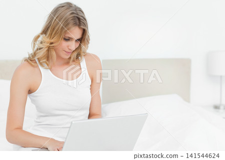 Woman in 20s sitting on bed wearing white tank top, using silver laptop by nightstand lamp Woman in 20s sitting on bed wearing white tank top, using silver laptop by nightstand lamp 141544624