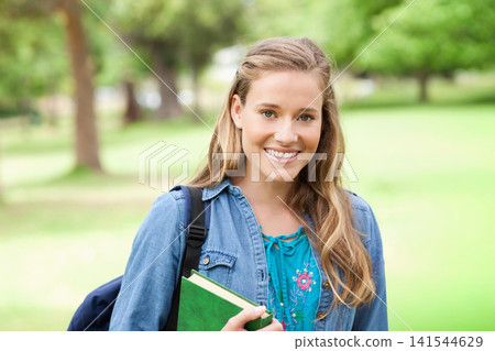Woman standing in park smiling, holding green hardcover notebook, wearing denim jacket and backpack Woman standing in park smiling, holding green hardcover notebook, wearing denim jacket and backpack 141544629