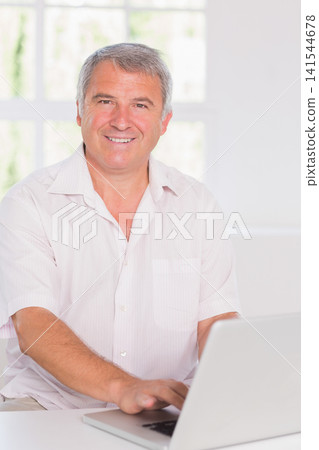 Mature male wearing short-sleeve shirt sitting at home using silver laptop by multi-pane window Mature male wearing short-sleeve shirt sitting at home using silver laptop by multi-pane window 141544678
