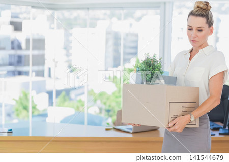 Woman in white blouse and skirt holding box with plant and papers at desk, copy space Woman in white blouse and skirt holding box with plant and papers at desk, copy space 141544679