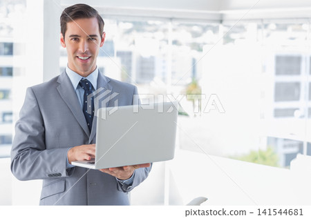 Man standing in modern office holding silver laptop, typing wearing light gray suit and navy tie Man standing in modern office holding silver laptop, typing wearing light gray suit and navy tie 141544681