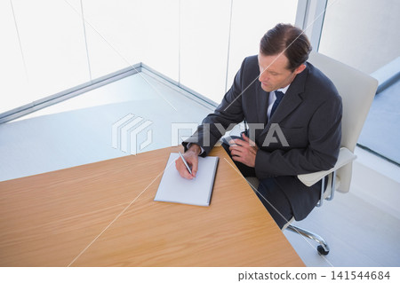 Mature man in dark suit writing with pen on spiral notebook at glass office, copy space 141544684