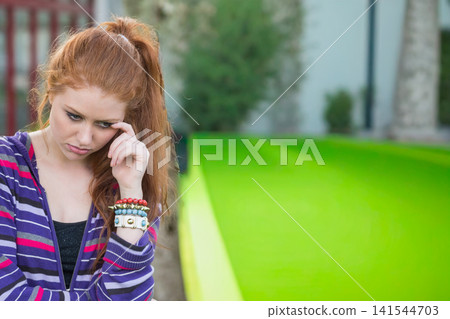 Female in 20s standing left on lawn rubbing temple, wearing purple hoodie and bracelets, copy space Female in 20s standing left on lawn rubbing temple, wearing purple hoodie and bracelets, copy space 141544703