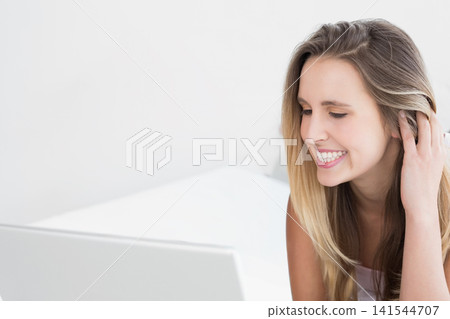 Woman in sleeveless top smiling while brushing hair leaning toward open laptop on bed, copy space Woman in sleeveless top smiling while brushing hair leaning toward open laptop on bed, copy space 141544707
