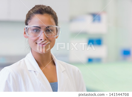 Female scientist wearing white lab coat and goggles, standing in clinical lab by bench and shelving Female scientist wearing white lab coat and goggles, standing in clinical lab by bench and shelving 141544708