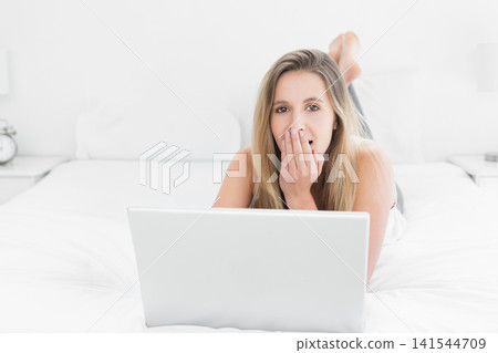 Woman 20s wearing white top lying on bed using silver laptop, covering mouth, alarm clock lamp 141544709
