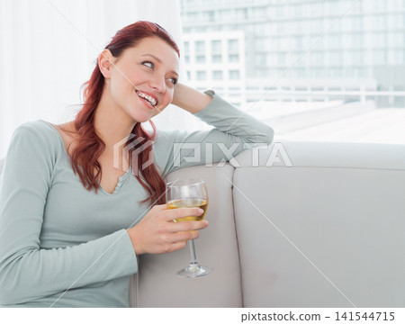 Female sitting on light-colored sofa near large windows wearing blue-gray top holding wine glass Female sitting on light-colored sofa near large windows wearing blue-gray top holding wine glass 141544715