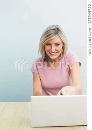 Woman smiling, pointing at silver laptop, working at studio light wood table in light pink T-shirt Woman smiling, pointing at silver laptop, working at studio light wood table in light pink T-shirt 141544720
