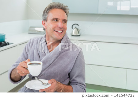 Mature man sitting at kitchen counter wearing gray robe, holding white cup and saucer with teaspoon Mature man sitting at kitchen counter wearing gray robe, holding white cup and saucer with teaspoon 141544722