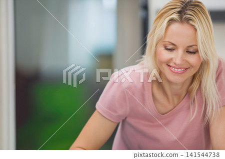 Middle-aged woman wearing pink T-shirt leaning forward smiling and looking down at home plant Middle-aged woman wearing pink T-shirt leaning forward smiling and looking down at home plant 141544738