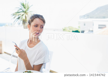 Woman sitting on sunny balcony wearing tee holding phone turning and looking worried at salad Woman sitting on sunny balcony wearing tee holding phone turning and looking worried at salad 141544742