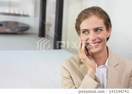 Woman sitting right in office wearing beige blazer holding phone to ear, smiling left, copy space Woman sitting right in office wearing beige blazer holding phone to ear, smiling left, copy space 141544743