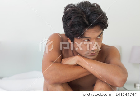 Mid adult Asian man sitting shirtless on bed, leaning forward and crossing arms, lamp in bedroom Mid adult Asian man sitting shirtless on bed, leaning forward and crossing arms, lamp in bedroom 141544744