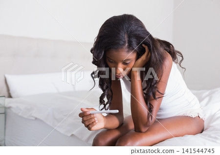African American female sitting on bed holding pregnancy test, resting head, wearing white tank top African American female sitting on bed holding pregnancy test, resting head, wearing white tank top 141544745