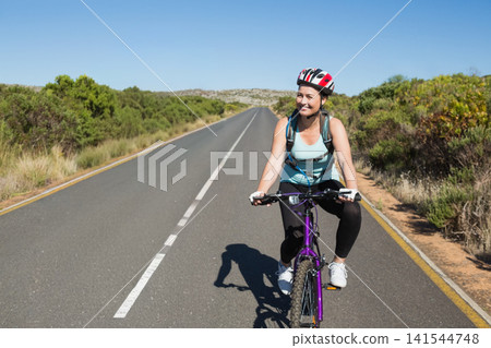 Female cyclist riding purple mountain-bike wearing helmet, gloves and pack on road with dashed-line Female cyclist riding purple mountain-bike wearing helmet, gloves and pack on road with dashed-line 141544748