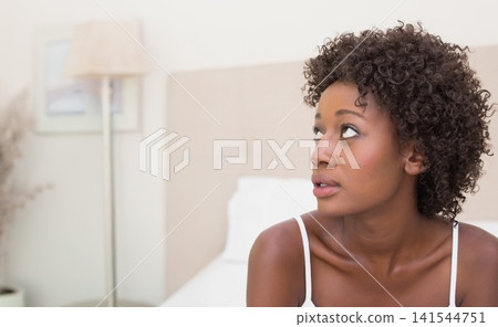 African American woman sitting on bed in bedroom in camisole by headboard, lamp, copy space African American woman sitting on bed in bedroom in camisole by headboard, lamp, copy space 141544751