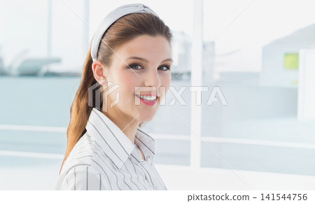 Woman 20s wearing striped shirt, headband smiling at camera at office windows with blurred rooftops 141544756