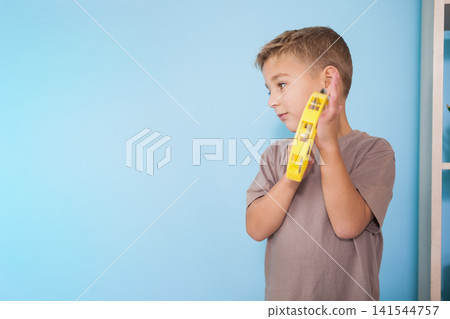 Child boy holding yellow tambourine to ear in room beside white shelf with plant, copy space Child boy holding yellow tambourine to ear in room beside white shelf with plant, copy space 141544757