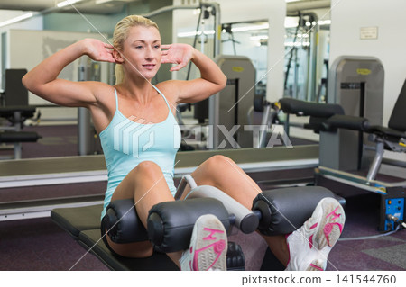 Fit female performing abdominal crunch on padded bench with rollers at gym, wearing blue tank top Fit female performing abdominal crunch on padded bench with rollers at gym, wearing blue tank top 141544760