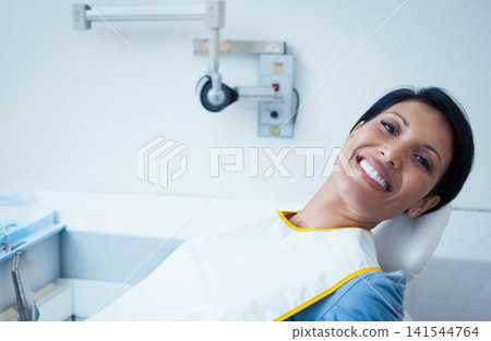 Female patient reclining in dental chair, wearing protective bib, showing teeth, tray and arm seen 141544764