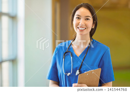 Asian female in blue scrubs standing in bright clinic corridor holding clipboard and stethoscope Asian female in blue scrubs standing in bright clinic corridor holding clipboard and stethoscope 141544768