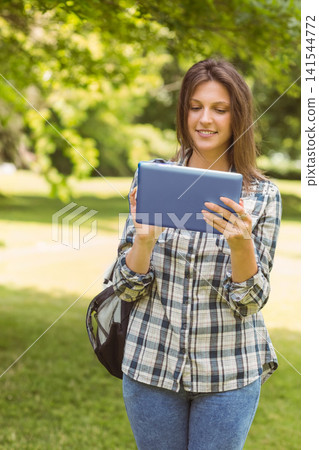 Female teenager standing on sunlit park lawn holding blue tablet, wearing plaid shirt and jeans Female teenager standing on sunlit park lawn holding blue tablet, wearing plaid shirt and jeans 141544772