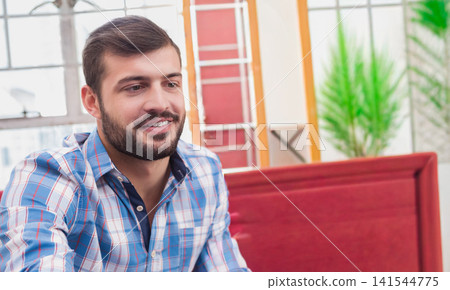 Bearded man sitting on red sofa in living room by window, plant, ladder, wearing plaid shirt Bearded man sitting on red sofa in living room by window, plant, ladder, wearing plaid shirt 141544775