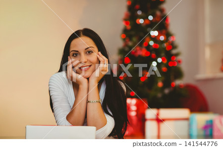 Woman reclining at home by laptop, wearing white long-sleeve top, smiling beside Christmas tree Woman reclining at home by laptop, wearing white long-sleeve top, smiling beside Christmas tree 141544776