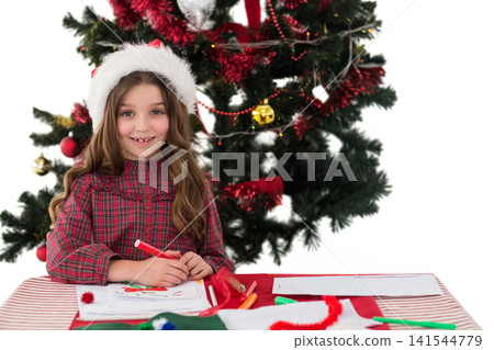 Child girl wearing Santa hat drawing and facing camera at craft table home near Christmas tree Child girl wearing Santa hat drawing and facing camera at craft table home near Christmas tree 141544779