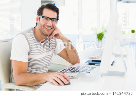 Man sitting at white desk in bright modern office reviewing printed thumbnails and using mouse 141544783