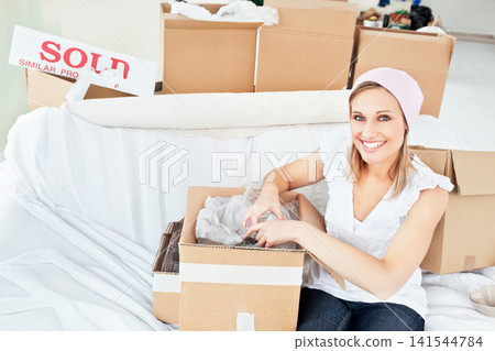 Woman sitting on living room floor unpacking bubble wrap from moving boxes, sold sign, copy space Woman sitting on living room floor unpacking bubble wrap from moving boxes, sold sign, copy space 141544784