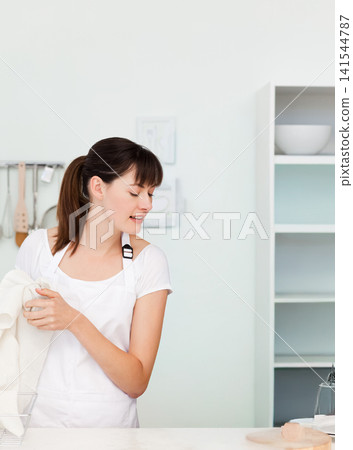 Woman wearing white apron standing at kitchen counter holding white dish towel near utensil rail Woman wearing white apron standing at kitchen counter holding white dish towel near utensil rail 141544787