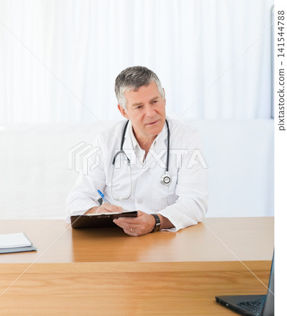 Senior male physician sitting at desk in clinic writing on clipboard, wearing lab coat, stethoscope Senior male physician sitting at desk in clinic writing on clipboard, wearing lab coat, stethoscope 141544788