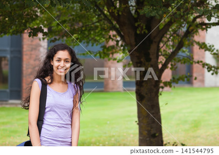 Woman standing facing camera on lawn by tree and brick building carrying black backpack, copy space Woman standing facing camera on lawn by tree and brick building carrying black backpack, copy space 141544795